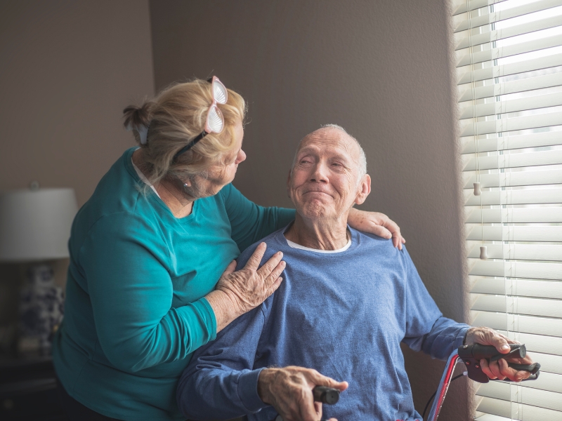 Caregiver talking and supporting elderly man during respite care visit.